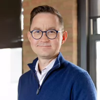 A professional headshot of a man wearing glasses and a navy blue sweater, standing in an office setting with natural light.