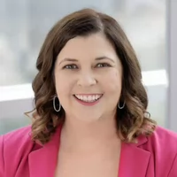 Professional headshot of a woman with curly hair wearing a pink blazer and earrings, smiling warmly.