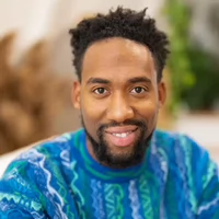 A smiling man with a stylish beard and curly hair, wearing a colorful knit sweater, seated in a cozy environment.