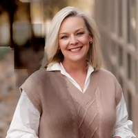 A smiling woman with shoulder-length blonde hair wearing a beige vest over a white shirt, standing in an outdoor setting.