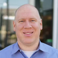 Headshot of a smiling man with a bald head, wearing a light blue shirt, suitable for professional contexts.