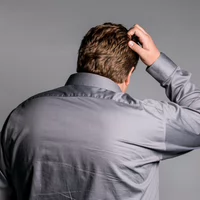 A man in a gray button-up shirt scratching his head, suggesting confusion or contemplation.