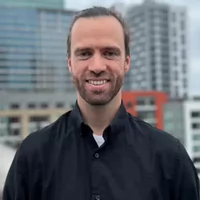 A smiling man in a black shirt stands against a modern cityscape background.