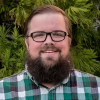 A smiling man with glasses and a beard wearing a green and black checkered shirt, standing in front of a lush green background.