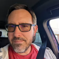 A close-up portrait of a man with glasses and a beard sitting in a car.